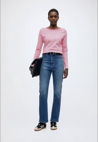 Ροζ και λευκή ριγέ μπλούζα με μακριά μανίκια, ψηλόμεσο μπλε τζιν, μαύρη ζώνη, κρατώντας μια μαύρη τσάντα, φορά μαύρα αθλητικά παπούτσια με λευκές λεπτομέρειες.