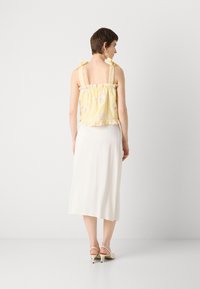Haut à motifs floraux jaunes avec un ourlet à volants et des bretelles en forme de nœud, associé à une jupe blanche lisse jusqu'au genou et des sandales à talons beiges.