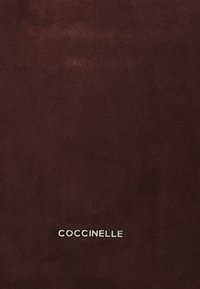 Brown suede material with a smooth texture, featuring a silver "COCCINELLE" logo at the bottom right corner. Minimalist design.