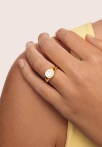 Anello in tono dorato caratterizzato da un design liscio e arrotondato con un emblema di faccia sorridente. La fascia è leggermente curvata e presenta una finitura lucida.