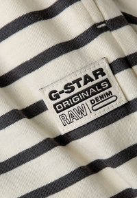 Tkanina G-Star w paski z kremowymi i ciemnoszarymi poziomymi liniami. Zawiera naszywkę z napisem „G-STAR ORIGINALS RAW DENIM” czarną czcionką.