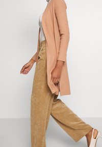 Rippstrick, langärmlige, pfirsichfarbene Strickjacke über einem figurbetonten weißen Top, kombiniert mit hellbraunen Cordstoff-Hose mit weitem Bein und beigen High Heels.