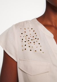 Haut beige à manches courtes, ornements dorés, noirs et violet clair disposés en motif dispersé sur l'épaule.