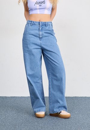 Person iført lyseblå Adidas crop top, højtaljede viddebenede blå jeans og beige sneakers, der står på gråt gulv.