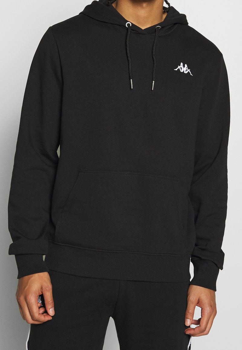 Personne portant un sweat à capuche noir avec un logo blanc représentant une silhouette double assise sur la poitrine et une poche kangourou devant.