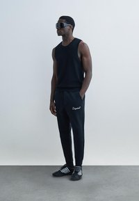 Mann iført svart ermeløs topp, svarte joggebukser med "Dsquared"-logo, store svarte solbriller og svarte joggesko med hvite striper.