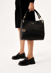 Una persona in abito nero tiene una borsa nera con texture e accenti dorati, indossando mocassini neri con plateau su uno sfondo bianco.