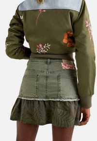 Chaqueta bomber verde oliva con bordados florales, combinada con una falda de mezclilla verde oliva que presenta un dobladillo deshilachado y un volante de capas con estampados.