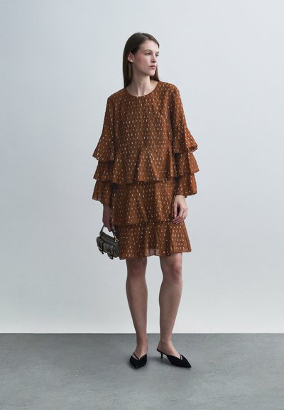 Robe brune à volants avec motifs blancs, manches longues à volants et encolure ronde. Associée à des chaussures noires à bouts pointus et un petit sac à main.