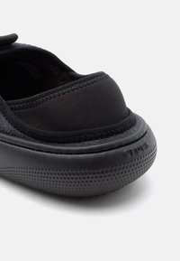 Prim-plan al unui pantof slip-on negru, cu toc și talpă din cauciuc texturat și partea superioară din țesătură, având marginile cusute și inscriptionarea „crocs” embosată.