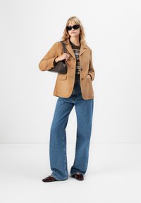 Kvinne iført beige blazer, mønstret genser, bredbeinte blå jeans, mørkerøde sko, svarte solbriller og bærende en brun håndveske over skulderen.