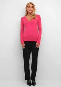 Roze gebreide trui met V-hals en lange mouwen, met knoopaccenten op de manchetten, gecombineerd met zwarte tailored broek en zwarte enkellaarsjes.