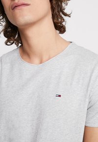 Szara bawełniana koszulka z okrągłym dekoltem i krótkimi rękawami. Posiada małą naszywkę z logo w kolorach czerwonym, białym i niebieskim po lewej stronie klatki piersiowej.