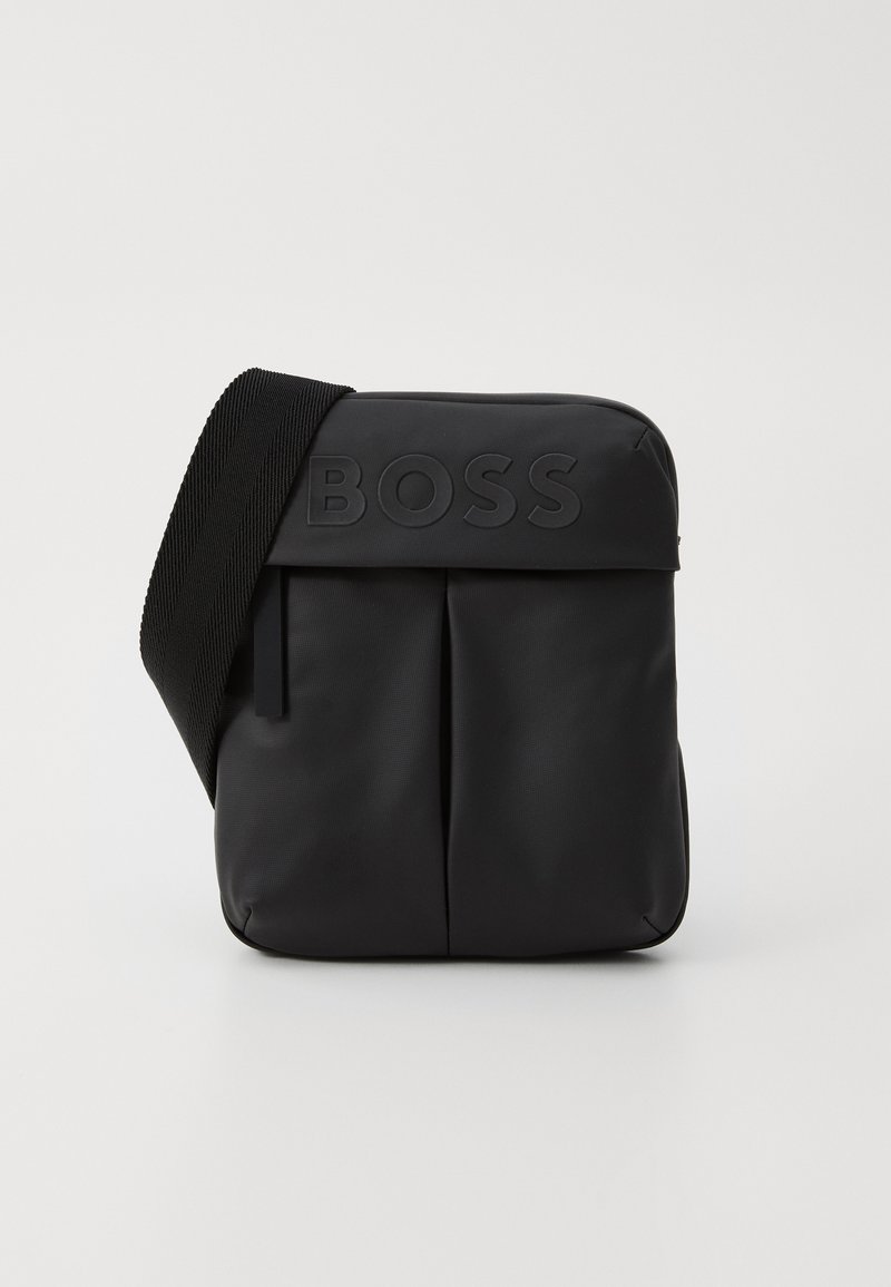 Sac bandoulière noir en matériau synthétique lisse. Dispose d'un rabat avant avec un logo en relief et d'une sangle tissée pour un transport facile.