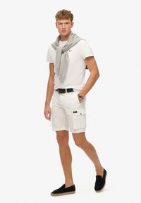 Pantalones cortos cargo blancos, camiseta blanca lisa, suéter gris sobre los hombros, espadrilles negras y cinturón negro con hebilla plateada.