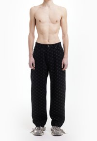 Pantalon noir avec un motif à logos, coupe décontractée et fermeture par bouton. Porté avec des baskets grises.