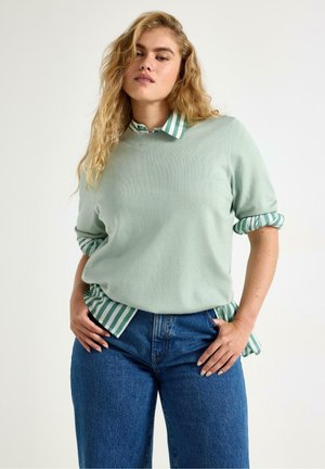 Kvinde med langt blondt hår iført en lysegrøn sweater over en grøn- og hvidstribet skjorte og blå jeans, stående med hænderne på hofterne.