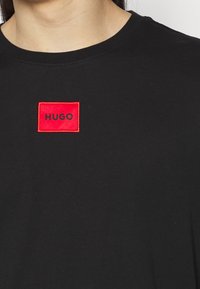 Person, der ein schwarzes T-Shirt mit einem kleinen roten Aufnäher auf der Brust trägt, auf dem in schwarzen Buchstaben "HUGO" steht.