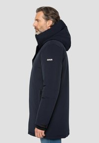 Lang marineblauw jas gemaakt van een textuurstof, met een grote capuchon en een logo op de mouw. Voorzien van ritsen en een getailleerd silhouet.