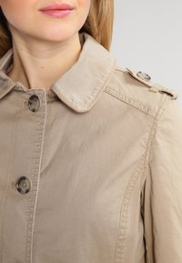 Beige trenchcoat met een knoopsluiting aan de voorkant, brede kraag en schouderlinten. Gemaakt van een gladde stof met zichtbare stikdetails.
