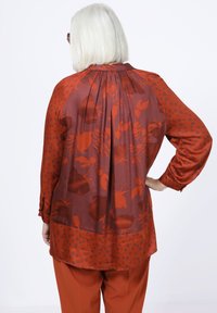 Langärmlige Bluse in Rostrot mit floralem und gepunktetem Muster. Glatte Textur mit Faltenlegung auf der Rückseite, abgerundetem Saum.