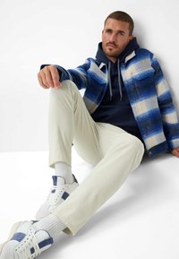 Giacca a quadri blu e bianca sopra un felpa blu navy, abbinata a pantaloni leggeri a coste e sneaker bianche con accenti blu.