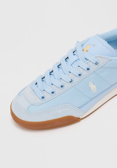 Sapatilhas em azul claro com um upper em lona texturizada e camurça, apresentando fecho de atacadores planos e um detalhe em logotipo dourado na lateral, além de uma sola em borracha.