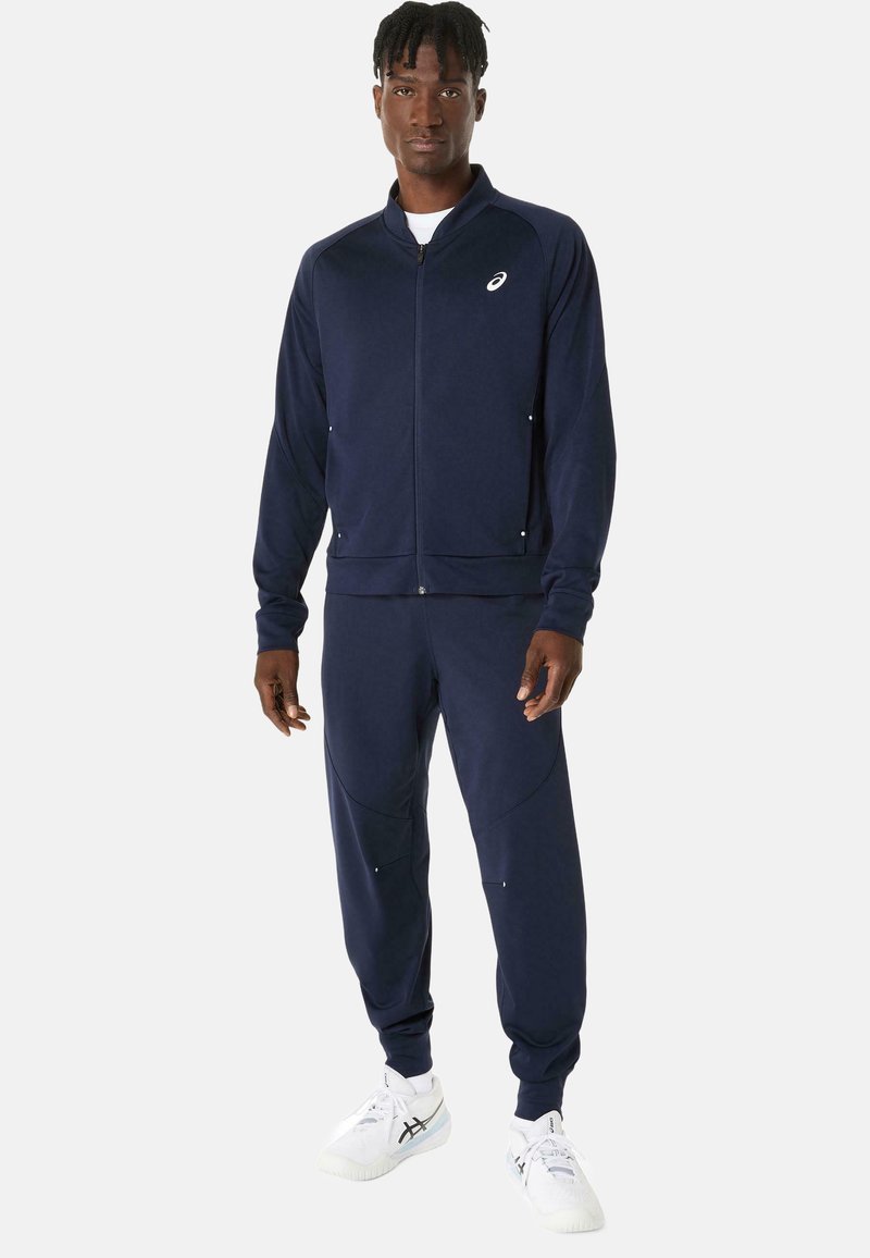 Tuta navy con giacca con zip e pantaloni. Tessuto morbido, design aderente con tasche laterali e logo sulla giacca. Sneakers bianche.