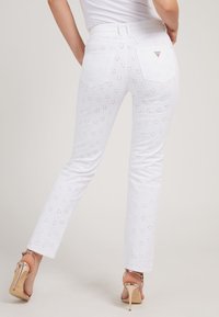 Pantaloni in denim bianco con motivi a fiori ritagliati, una silhouette aderente e due tasche posteriori. Abbinati a sandali con tacco alto color oro rosa.