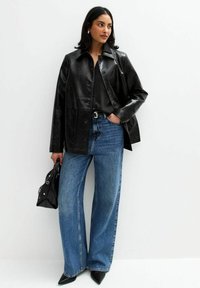 Chaqueta de cuero negra con cierre de botón, combinada con jeans de mezclilla azul de pierna ancha. Un bolso negro y tacones negros de punta completan el look.