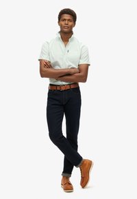 Camicia a maniche corte di colore verde chiaro, jeans blu scuro, cintura di pelle marrone e stivali marroni. Il modello posa con le braccia incrociate su uno sfondo bianco.