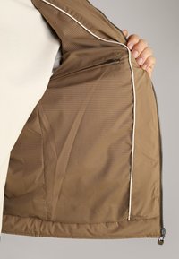 Interno di giacca marrone con fodera testurizzata, tasca con zip e piping di colore chiaro lungo i bordi. I materiali sembrano lisci e durevoli.