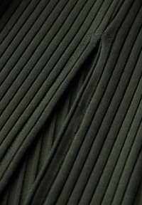 Tessuto a coste verde scuro con strisce verticali e una superficie morbida, con cuciture visibili e una leggera elasticità. Un dettaglio chiave è uno spacco.