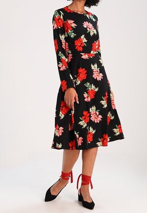 Schwarzes langärmliges Kleid mit rotem und rosa Blumenmuster, tailliertem Oberteil, ausgestelltem Rock, kombiniert mit schwarzen Absätzen mit roten Knöchelriemchen.