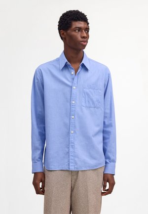 Giovane uomo che indossa una camicia celeste con bottoni e taschino sul petto, e pantaloni beige con texture, in piedi davanti a uno sfondo bianco semplice.