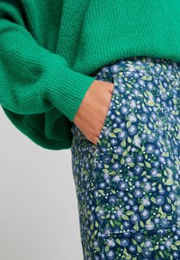 Jersey de punto verde combinado con pantalones de flores azules que tienen bolsillos. La tela presenta un aspecto texturizado con estampados vibrantes y coloridos.