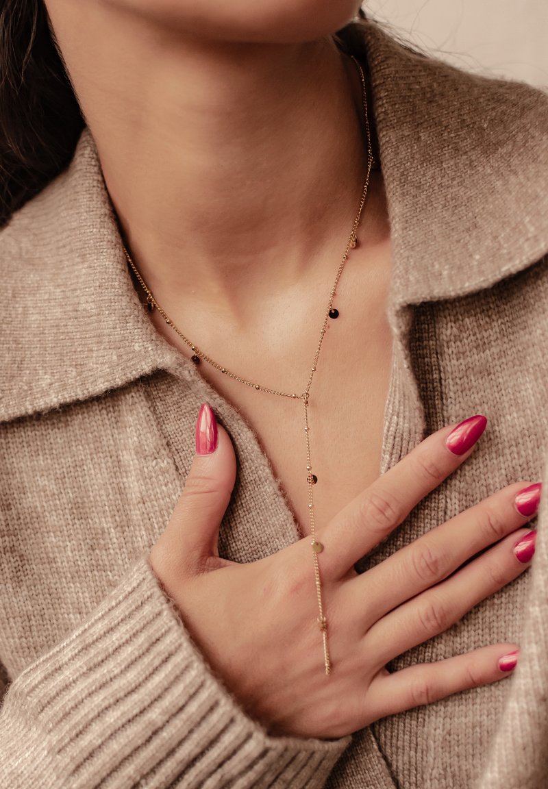 Collier en chaîne dorée avec de petits accents ronds, porté sur un pull beige clair. Les cercles sont noirs et dorés, avec un design délicat et allongé.