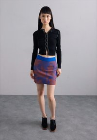 Cardigan lung cu mâneci lungi, de culoare neagră, cu nasturi, purtat cu o fustă modelată în nuanțe de purpuriu și albastru. Se evidențiază materialele texturate și combinațiile de culori unice.