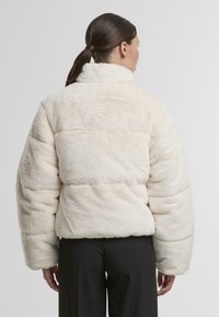 Veste en duvet de couleur crème avec texture douce, col haut et manches larges. Présente des coutures horizontales et une silhouette courte.