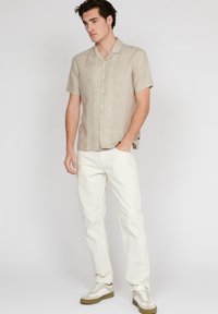 Camicia in lino a maniche corte di un beige chiaro con una vestibilità relax, abbinata a pantaloni bianchi e sneakers casual con dettagli color nocciola.