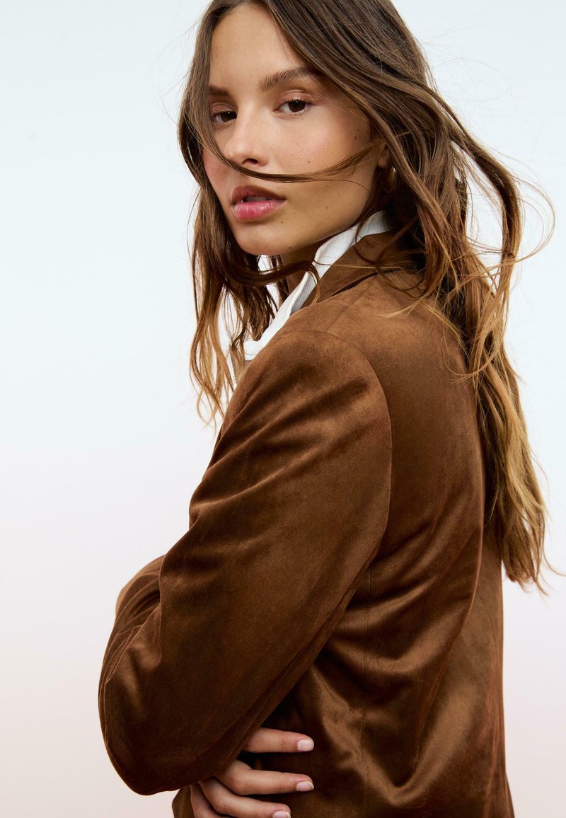 Blazer en suède marron au design ajusté, avec des revers crantés et un subtil éclat, porté sur une chemise blanche et avec des cheveux longs et lâches.