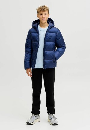 Marineblå pufferjakke med hætte, der har en blank overflade og vandret kviltet design. Båret over en lyseblå sweater og sorte bukser.