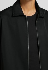 Czarna lekka kurtka z kołnierzem typu stójka, zamkiem błyskawicznym na całej długości i gładką fakturą. Założona na dopasowaną czarną bluzkę.