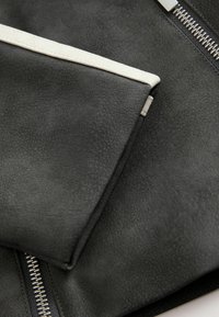 Svart läderplånbok med texturerad yta, med ett vitt accentband och silverfärgad dragkedja på sidan. Minimalistisk design.