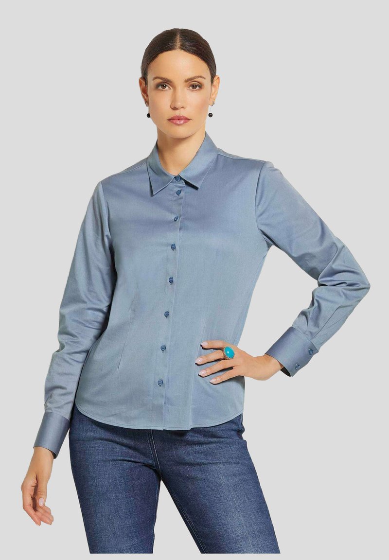 Himmelblaues, langärmeliges Hemd mit Knöpfen und Kragen, glatte Textur und passende blaue Knöpfe. Getragen mit dunklen Jeans und einem türkisfarbenen Ring.