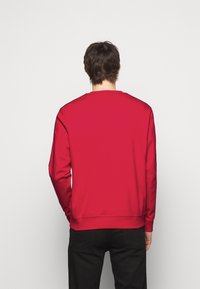 Felpa rossa con maniche lunghe e scollo rotondo. Texture liscia, polsini e orlo a coste, design semplice, senza loghi o motivi visibili.