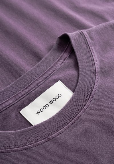 Lilla bomulls t-skjorte med rund hals, med en hvit merkelapp hvor "WOOD WOOD" er trykt i svart. Glatt tekstur og minimalistisk design.