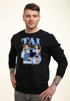 Mężczyzna w czarnej bluzie z kolorową grafiką trzech kobiet oraz dużym niebieskim napisem „TAI” na przodzie, stoi z rękami na biodrach.