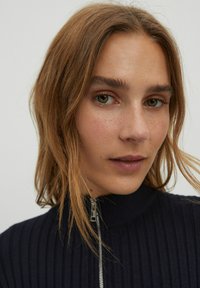 Marineblaues Rippstrick-Oberteil mit hohem Kragen und silbernem Reißverschluss, zu sehen ist eine Nahaufnahme des Gesichts einer Person mit schulterlangem Haar.