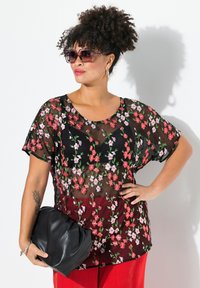 Top trasparente nero con ricami floreali in rosso e rosa; maniche corte; indossato sopra pantaloni rossi a coste; modella che tiene una borsa a mano nera.